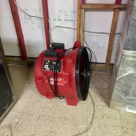 Red air mover running in a room where the lower wall has been cut open and plastic sheeting hangs after a leak at floor level
