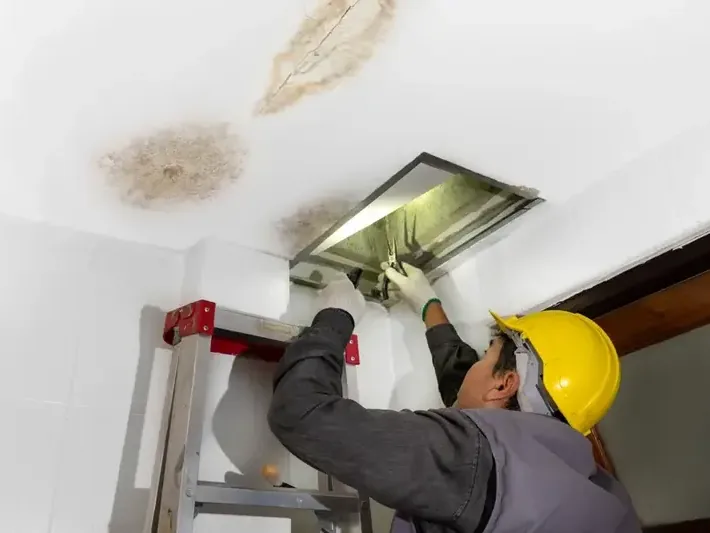 Worker checking an attic hatch beneath a water-stained ceiling from a roof leak